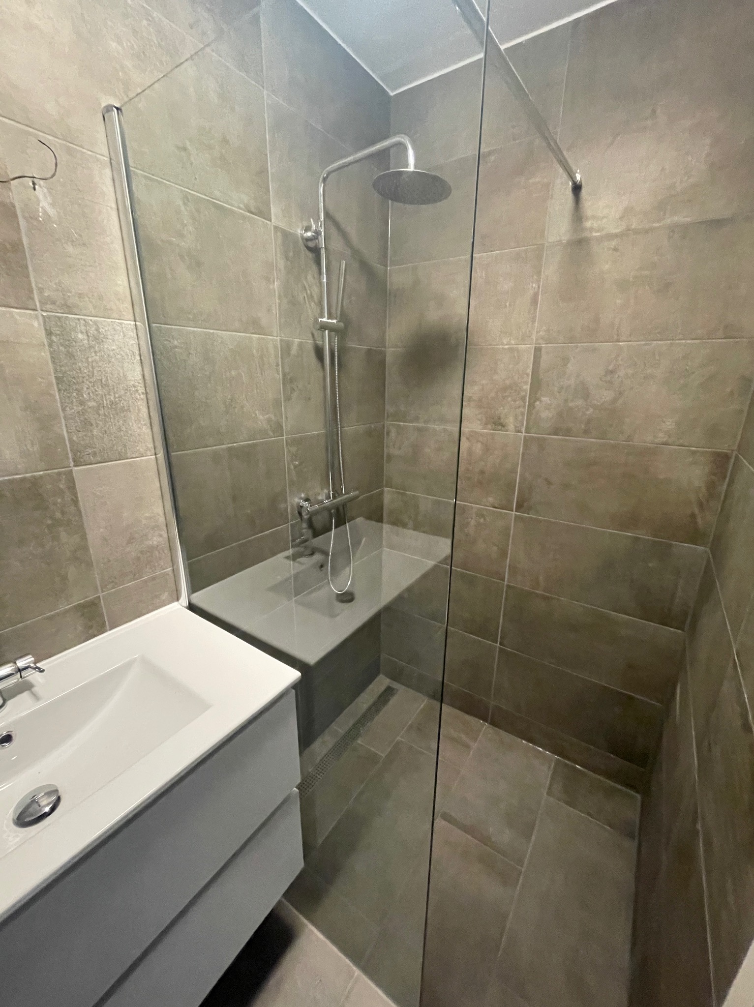 Nowoczesna kabina prysznicowa z deszczownicą i szklaną ścianką, obok umywalka z szafką w minimalistycznej łazience wykończonej płytkami w odcieniach szarości.