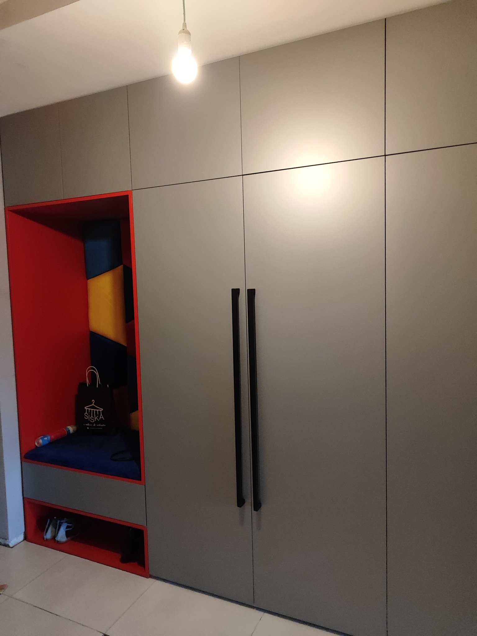 Szara szafa wnękowa z czerwonym regałem i siedziskiem, widoczna torba z logo firmy i buty na półce.