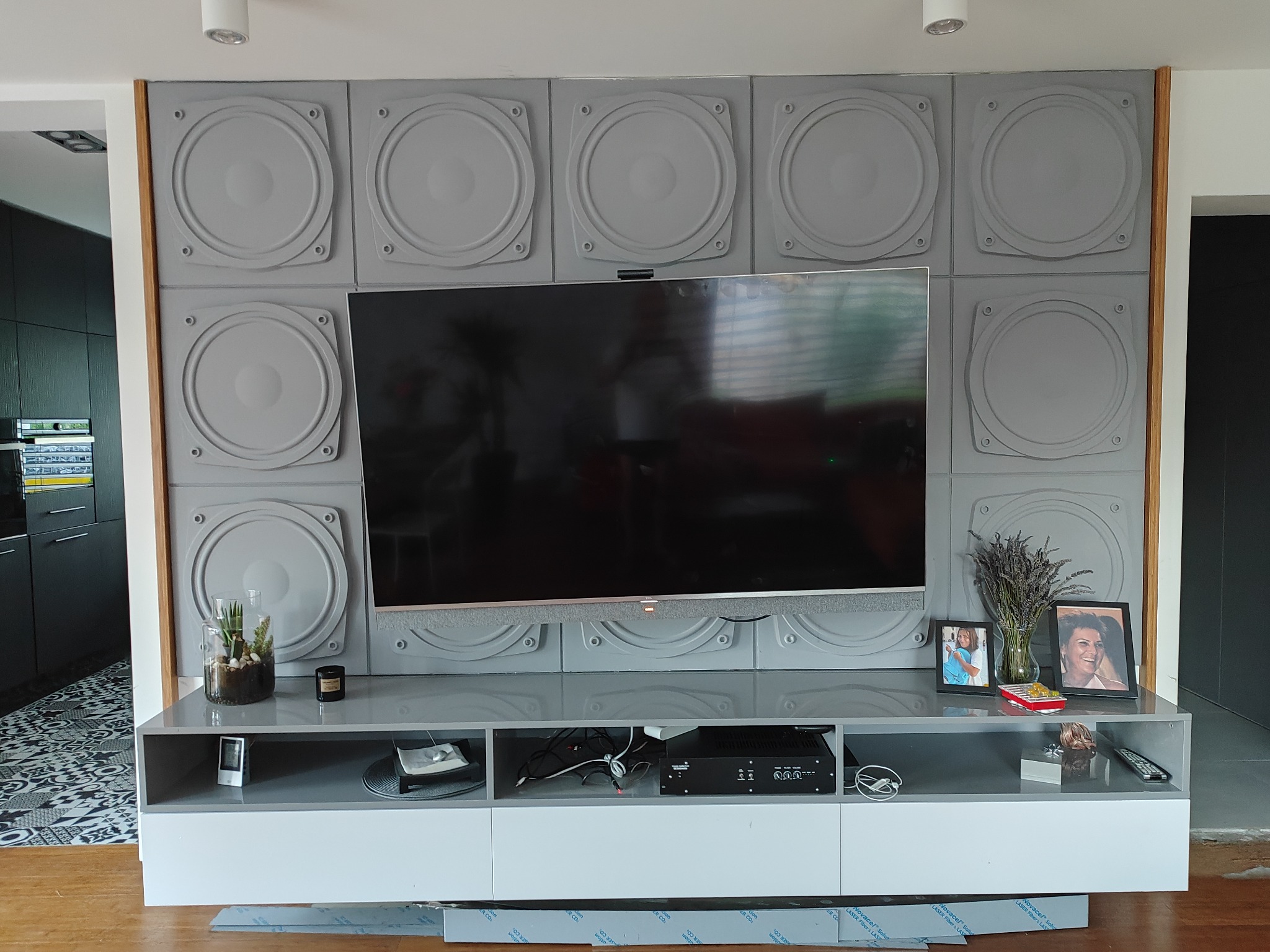 Szaro-biała szafka RTV z telewizorem i dekoracyjną ścianą z panelami w kształcie głośników, z widocznymi kablami i akcesoriami audio.