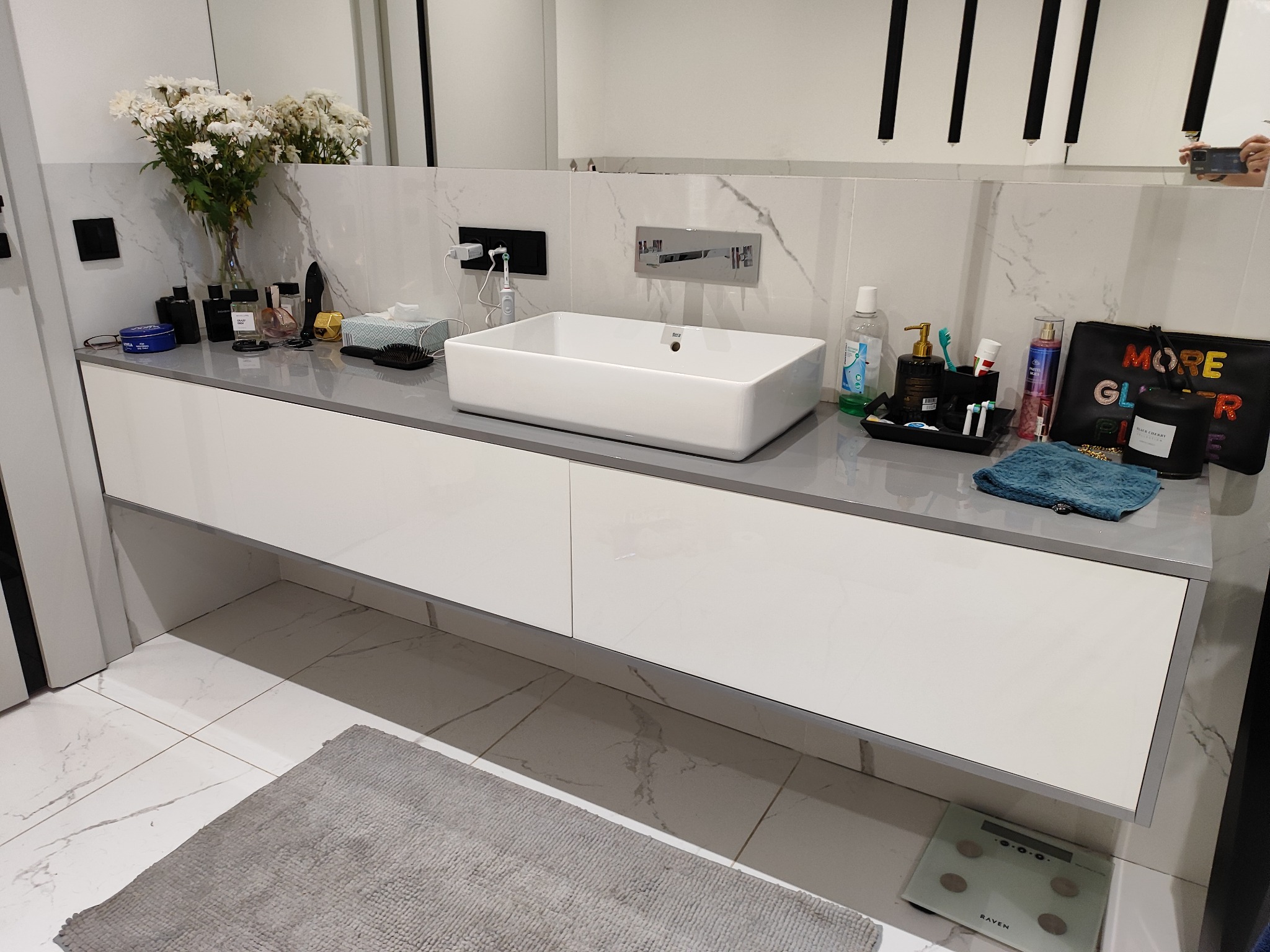 Nowoczesna łazienka z białą szafką pod umywalką z szarym blatem, na którym stoją kwiaty, kosmetyki i akcesoria łazienkowe, marmurowe płytki na ścianie i podłodze, w tle widoczne odbicie osoby...