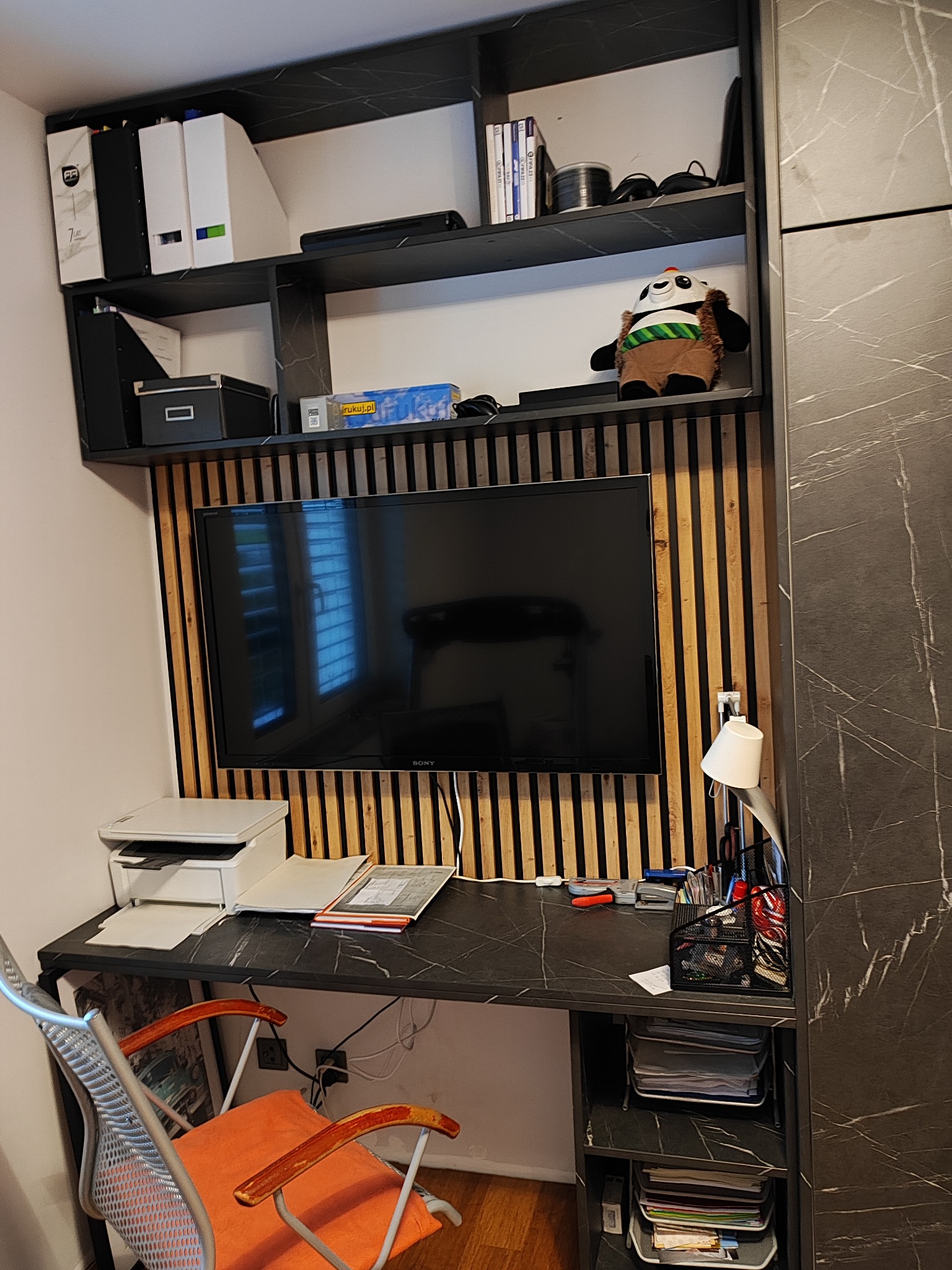 Czarno-białe biurko z marmurkowym blatem i drewnianą ścianką działową za telewizorem, uzupełnione półkami z książkami, ozdobami i pluszową pandą.