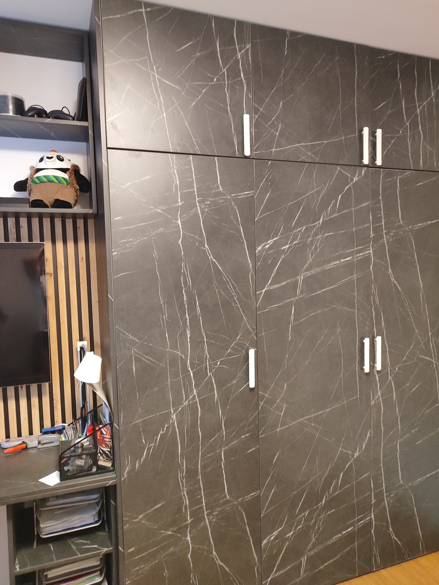 Duża, szara szafa z frontami imitującymi marmur, z podłużnymi, białymi uchwytami, przylegająca do biurka z półkami i regałem, na których stoją akcesoria biurowe i pluszowy miś.