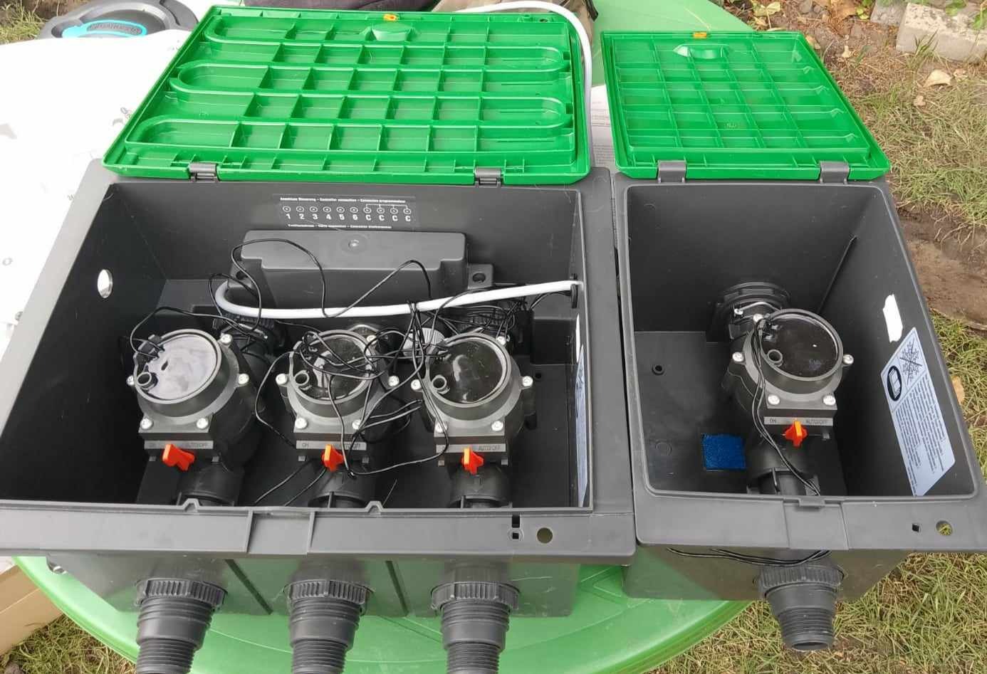 Rozdzielacz wody do systemu nawadniania w szarej skrzynce z zielonymi pokrywami, widoczne zawory z pomarańczowymi przełącznikami i okablowanie.