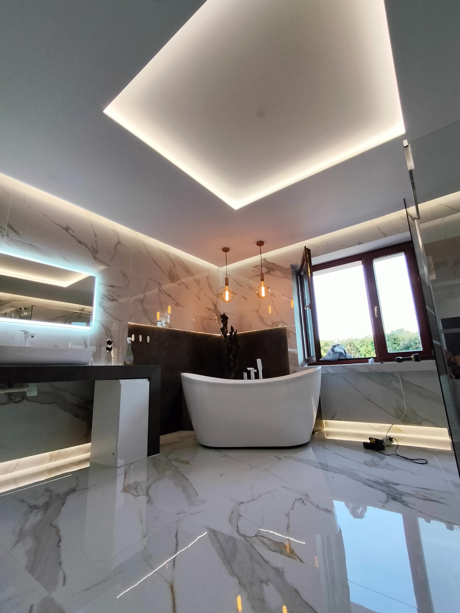 Nowoczesna łazienka z wolnostojącą wanną, marmurowymi płytkami na ścianach i podłodze, oświetleniem LED w suficie i pod wanną, oraz dekoracyjnymi lampami wiszącymi.