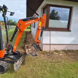 Pomarańczowa minikoparka Hitachi wykonuje wykop liniowy wzdłuż ściany budynku, widoczna ziemia w łyżce i wąski wykop w trawie, operator w kamizelce odblaskowej w kabinie.