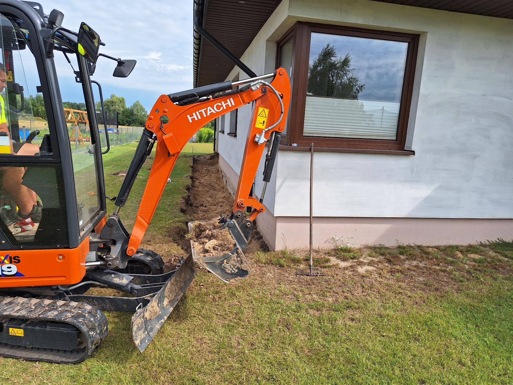 Pomarańczowa minikoparka Hitachi wykonuje wykop liniowy wzdłuż ściany budynku, widoczna ziemia w łyżce i wąski wykop w trawie, operator w kamizelce odblaskowej w kabinie.