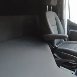 Wnętrze furgonetki z nową, szarą tapicerką na ścianach i siedzeniach, widok z boku na siedzenie kierowcy z zagłówkiem i podłokietnikiem.