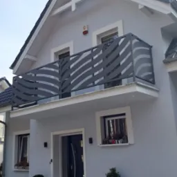 Nowoczesna balustrada balkonowa ze stali nierdzewnej z ażurowym wzorem falistych linii, zamontowana na balkonie domu w jasnych kolorach.