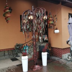 Kowalstwo Artystyczne i Metaloplastyka - Ozdobne metalowe drzewko z lampkami w formie wierzby płaczącej, stojące na zewnątrz przed domem z pomarańczową elewacją, obok białe donice z wrzosami i wiszące spiralne dekoracje.