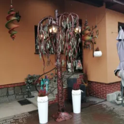 Ozdobne metalowe drzewko z lampkami w formie wierzby płaczącej, stojące na zewnątrz przed domem z pomarańczową elewacją, obok białe donice z wrzosami i wiszące spiralne dekoracje.