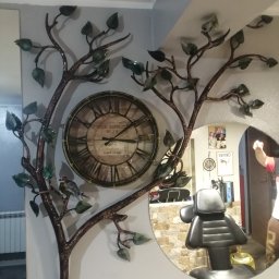 Kowalstwo Artystyczne i Metaloplastyka - Dekoracja ścienna z metalu: drzewo z zegarem i ptaszkiem, odbicie fotela w okrągłym lustrze w salonie, widoczne zdjęcia i plakaty na ścianie.