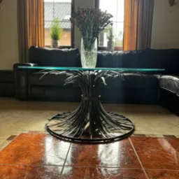 Szklany stolik kawowy z metalową podstawą w kształcie snopka zboża, na tle skórzanej sofy i okien z zasłonami, na błyszczącej, wzorzystej podłodze.