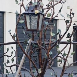 Kowalstwo Artystyczne i Metaloplastyka - Kuta, metalowa konstrukcja przypominająca drzewo z liśćmi i latarnią w stylu retro, na tle budynku z szarymi bramami garażowymi.