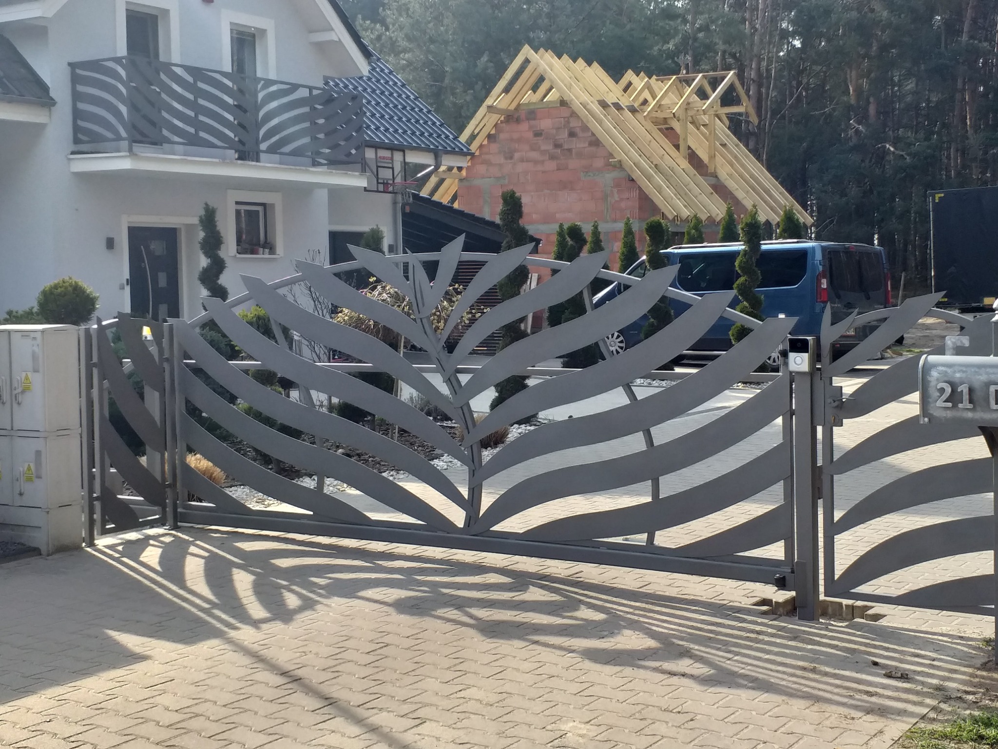 Otwarta brama wjazdowa z dekoracyjnym wzorem liścia na posesji z nowym domem w budowie i spiralnymi krzewami.