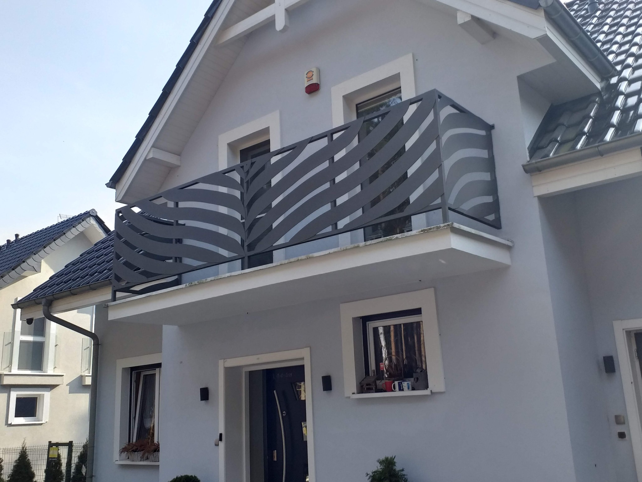 Nowoczesna balustrada balkonowa ze stali nierdzewnej z ażurowym wzorem falistych linii, zamontowana na balkonie domu w jasnych kolorach.