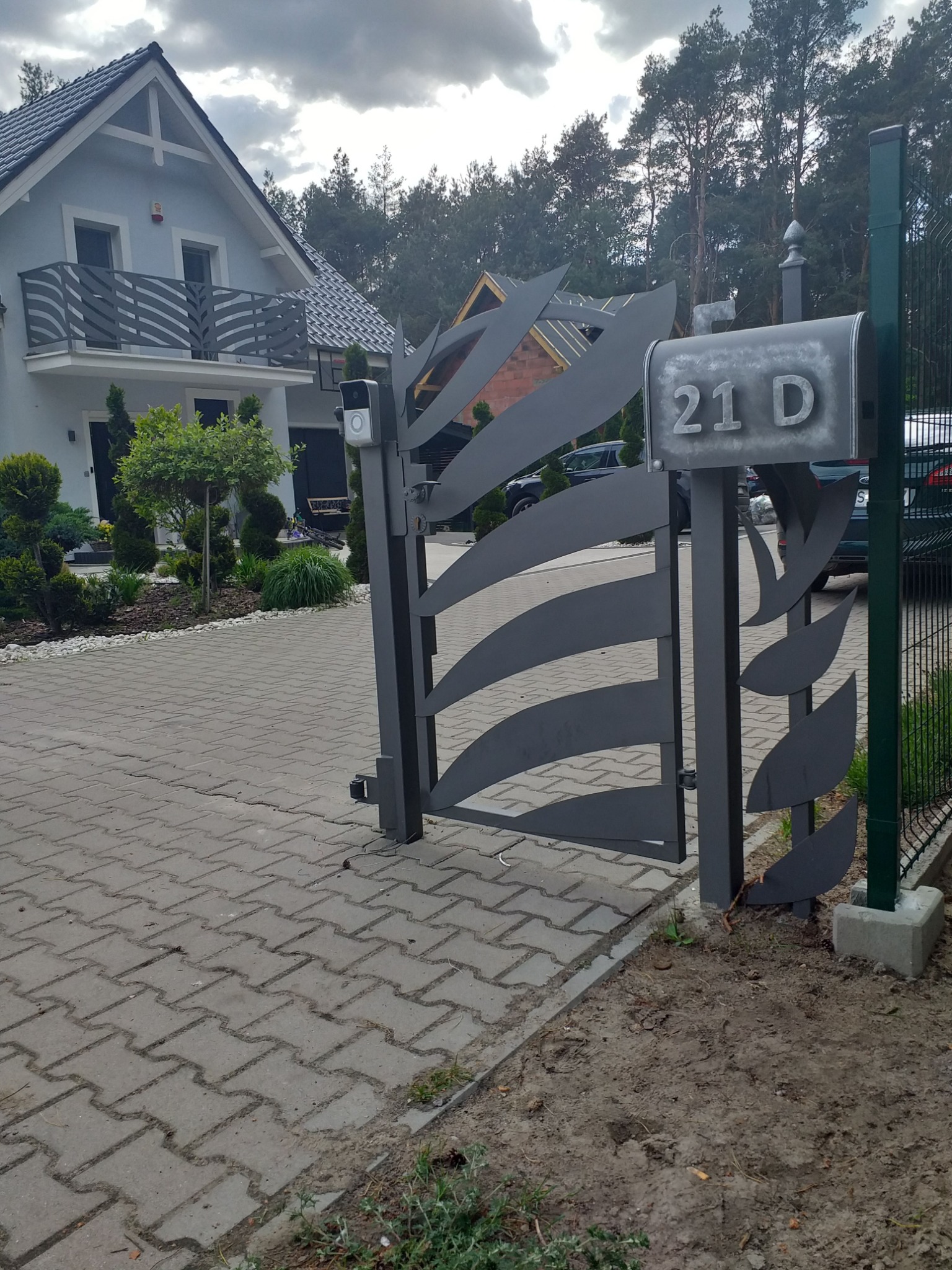 Nowoczesna, metalowa brama wjazdowa z motywem liści, szara skrzynka pocztowa z numerem 21D, na tle domu z balkonem i zadbanego ogrodu.