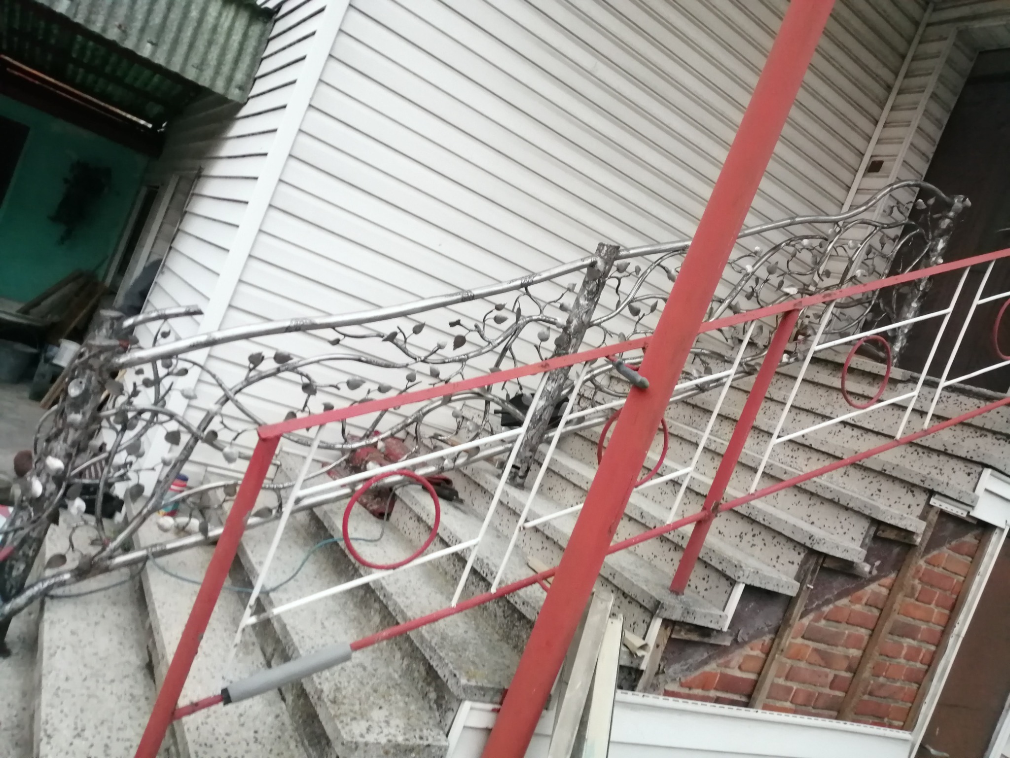 Schody zewnętrzne z metalową balustradą o dekoracyjnym wzorze liści, czerwone elementy konstrukcyjne, widoczne niedokończone prace i materiały budowlane w tle.