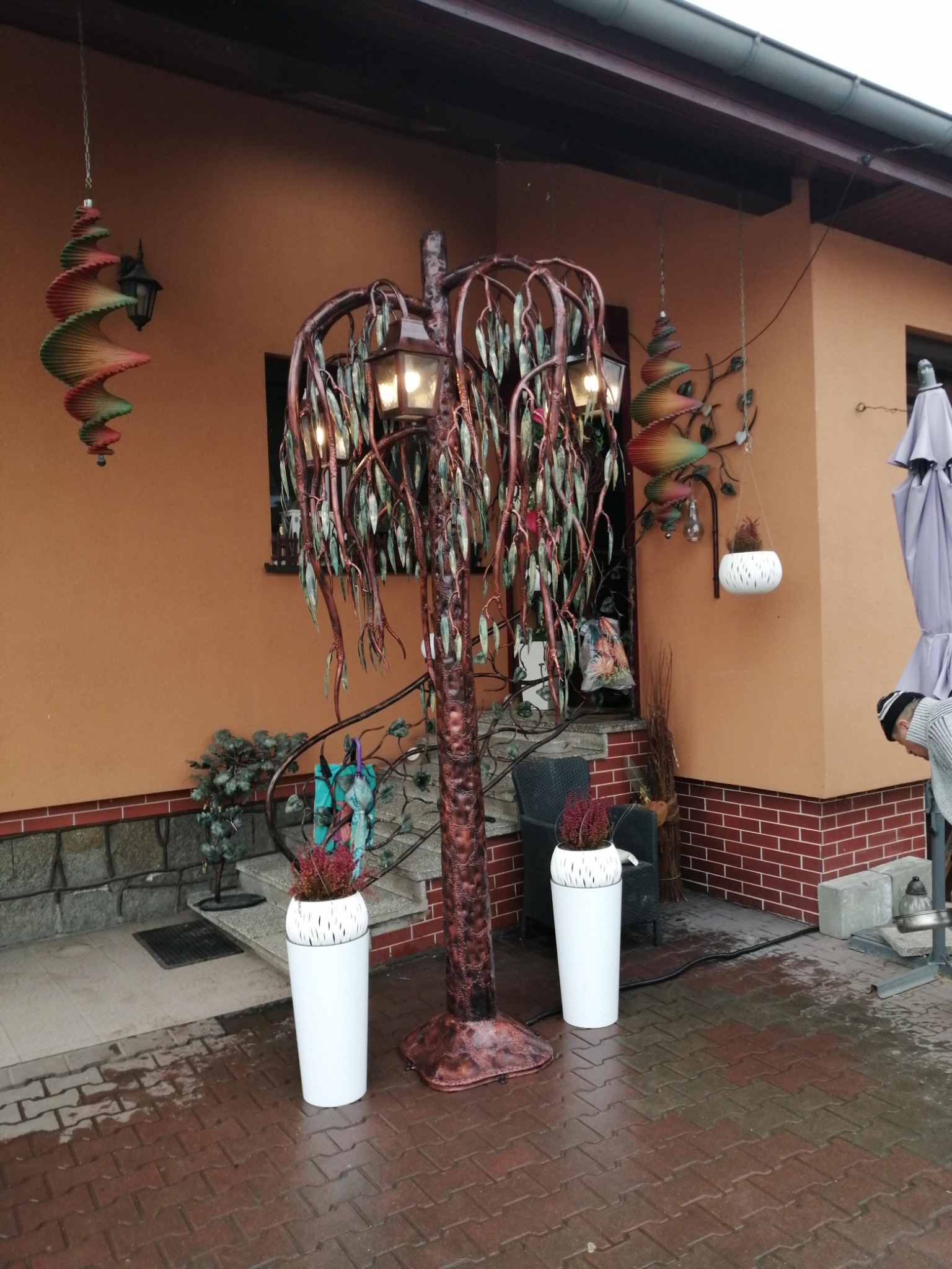 Ozdobne metalowe drzewko z lampkami w formie wierzby płaczącej, stojące na zewnątrz przed domem z pomarańczową elewacją, obok białe donice z wrzosami i wiszące spiralne dekoracje.