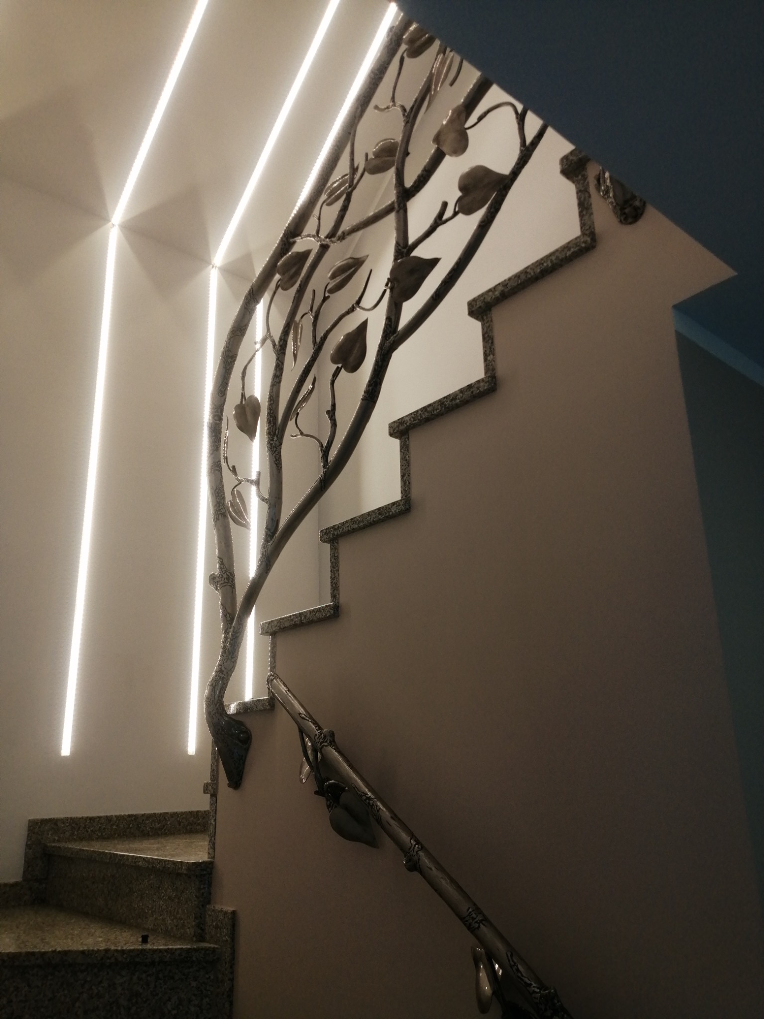 Schody z metalową balustradą w kształcie drzewa z liśćmi, oświetlone paskami LED na ścianie, widoczne marmurowe stopnie.