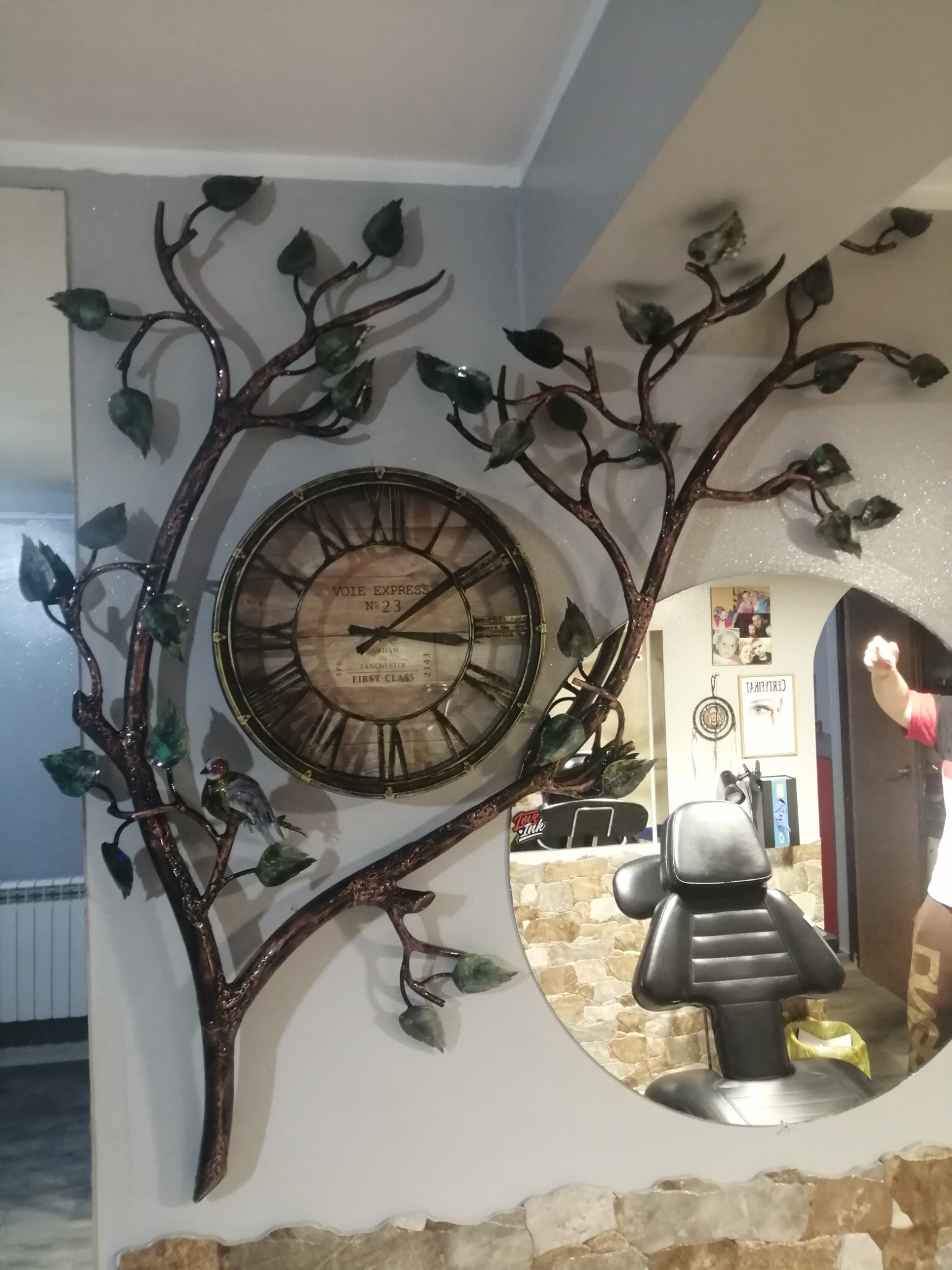 Dekoracja ścienna z metalu: drzewo z zegarem i ptaszkiem, odbicie fotela w okrągłym lustrze w salonie, widoczne zdjęcia i plakaty na ścianie.