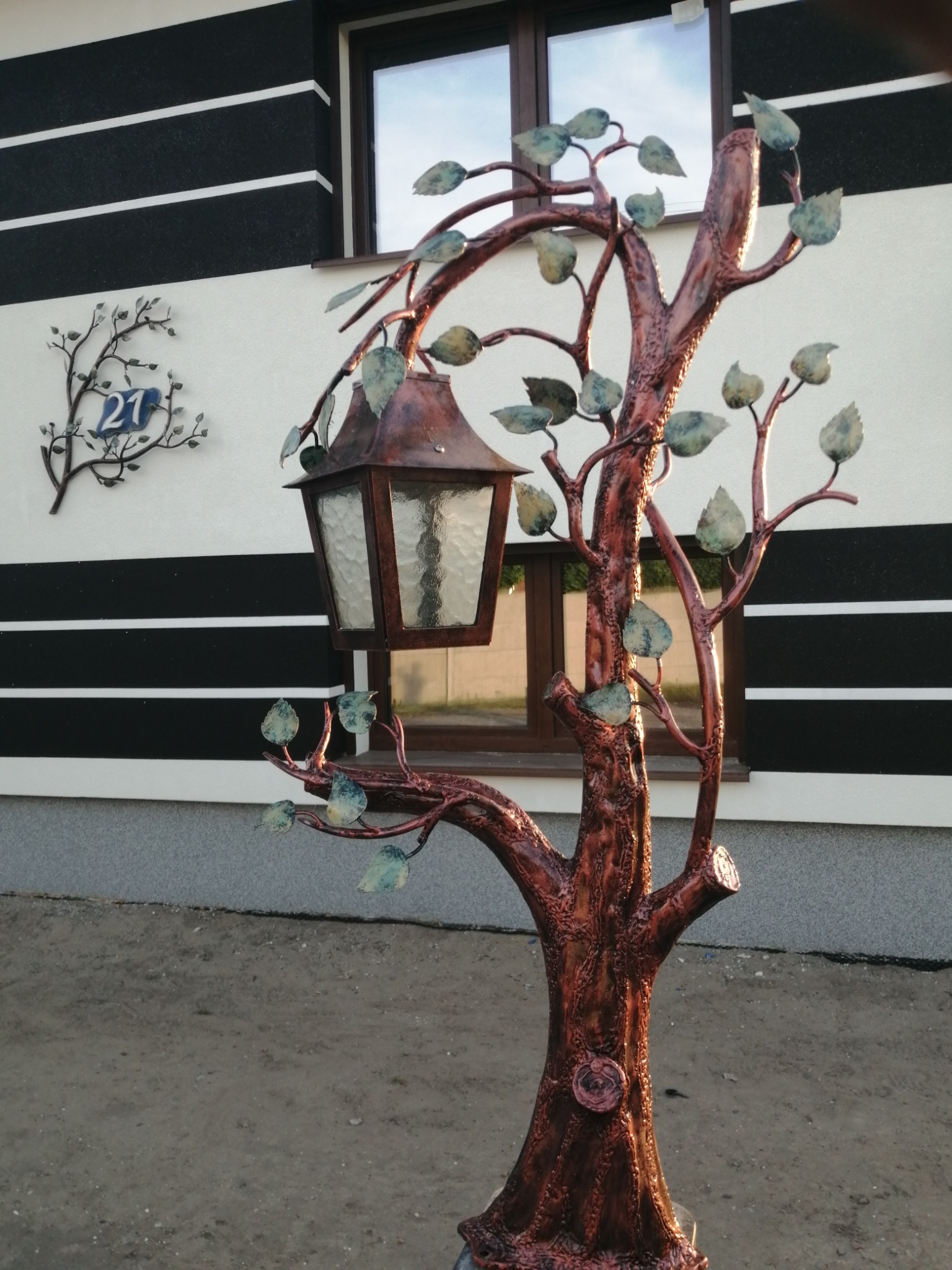 Kuta, metalowa lampa ogrodowa w kształcie drzewa z patynowanymi liśćmi, na tle elewacji budynku z dekoracyjnymi pasami i numerem domu.