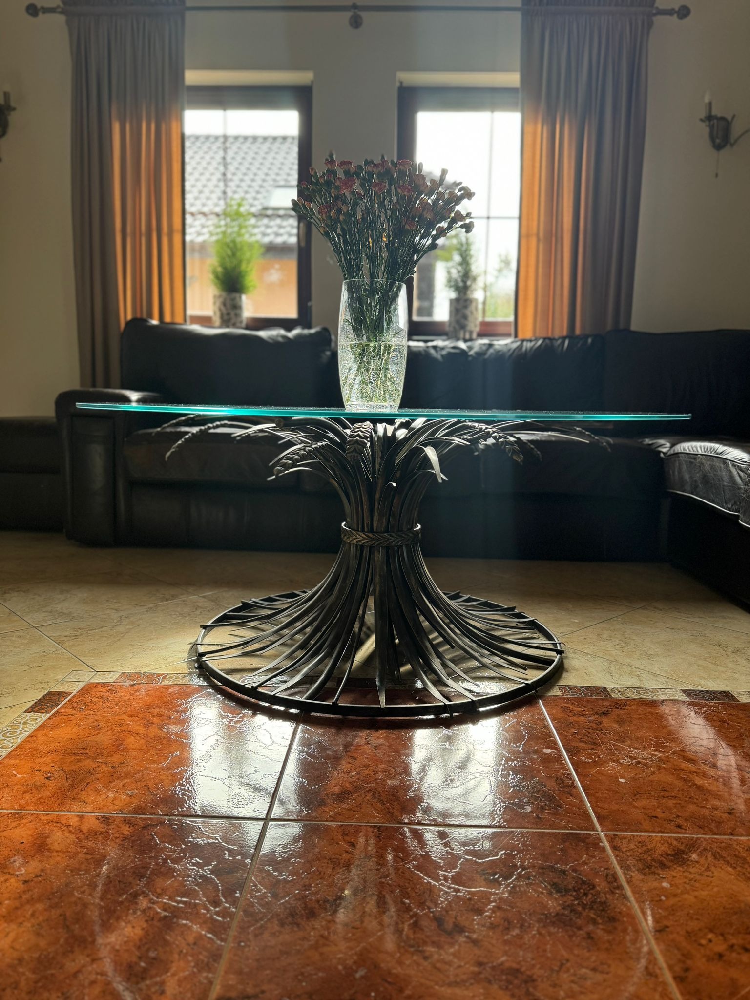 Szklany stolik kawowy z metalową podstawą w kształcie snopka zboża, na tle skórzanej sofy i okien z zasłonami, na błyszczącej, wzorzystej podłodze.