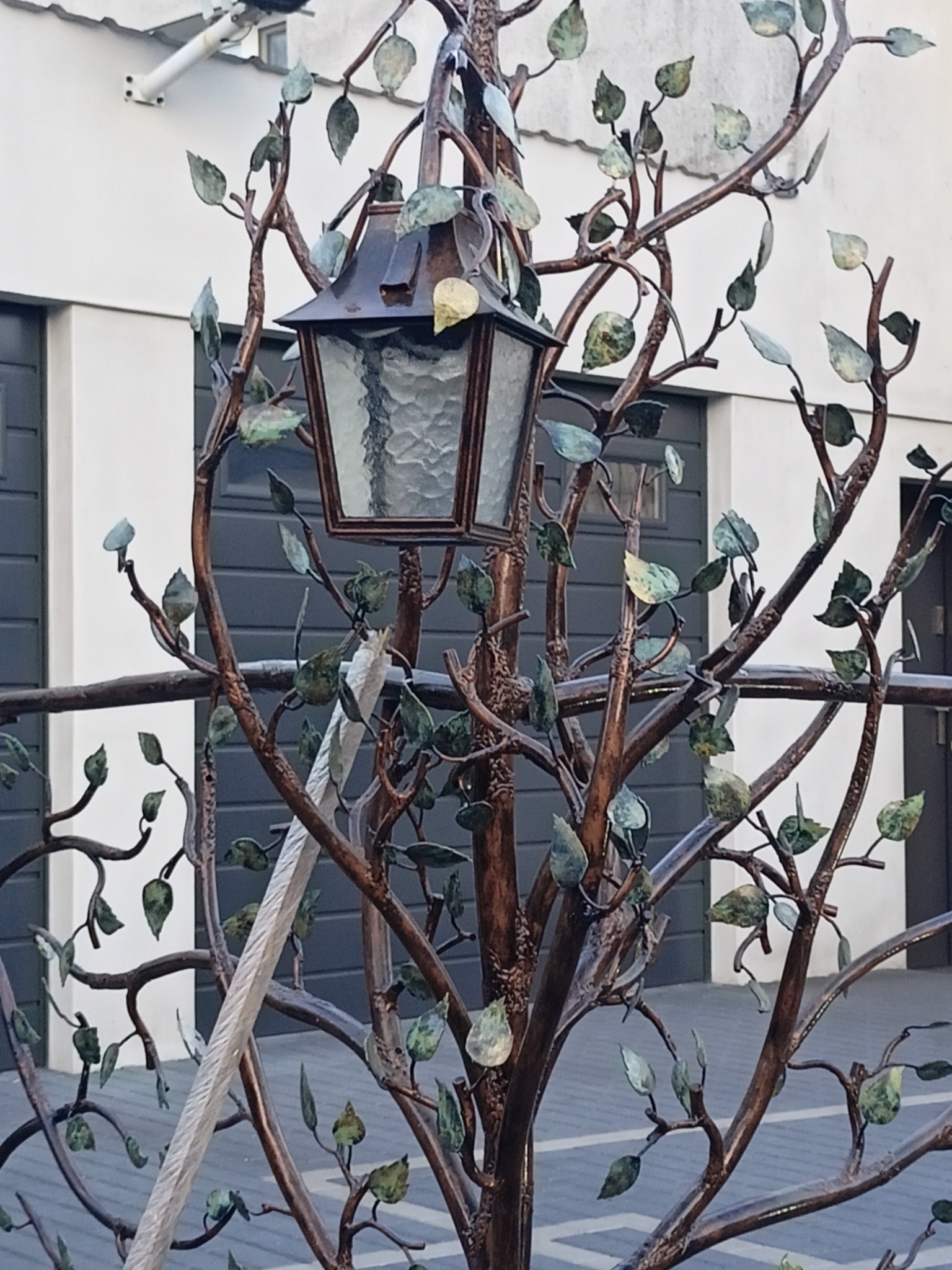 Kuta, metalowa konstrukcja przypominająca drzewo z liśćmi i latarnią w stylu retro, na tle budynku z szarymi bramami garażowymi.