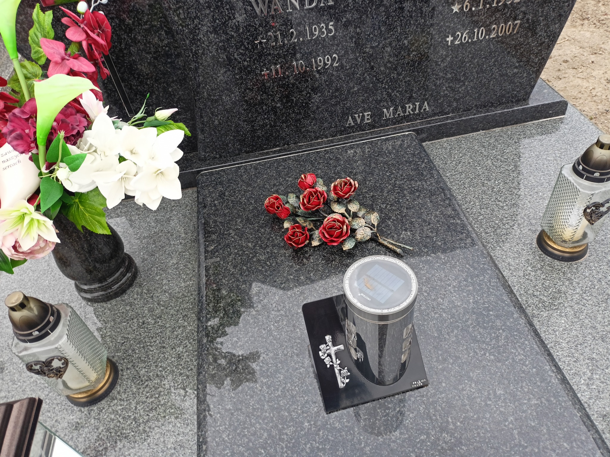Granitowy nagrobek z wyrytymi imionami i datami, ozdobiony wiązanką czerwonych róż, wazonem z kolorowymi kwiatami i lampionami, w tym jednym solarnym na czarnej podstawce z krzyżem.