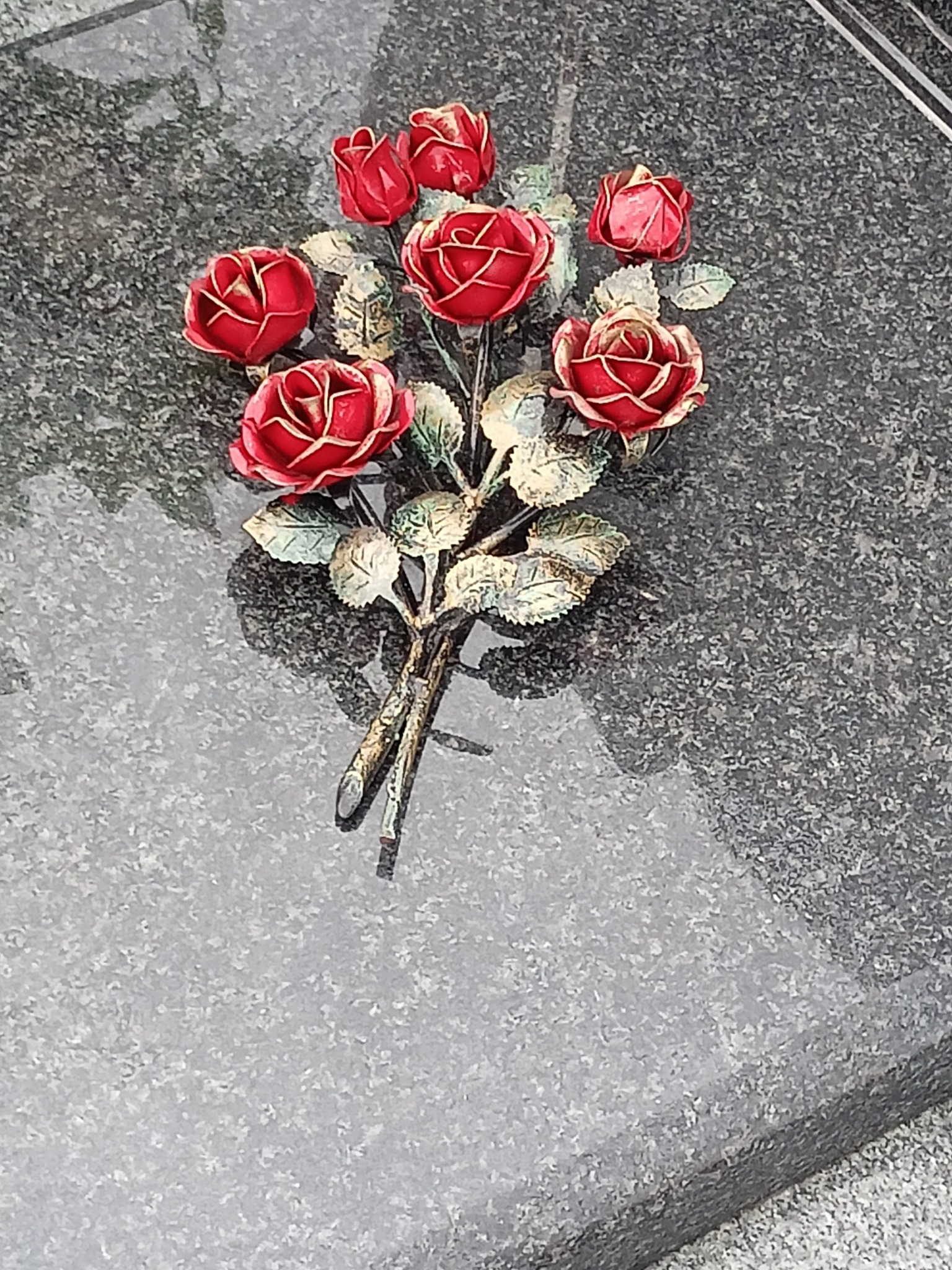 Metalowy bukiet czerwonych róż z zielonymi listkami na szarym, wypolerowanym nagrobku z granitu.