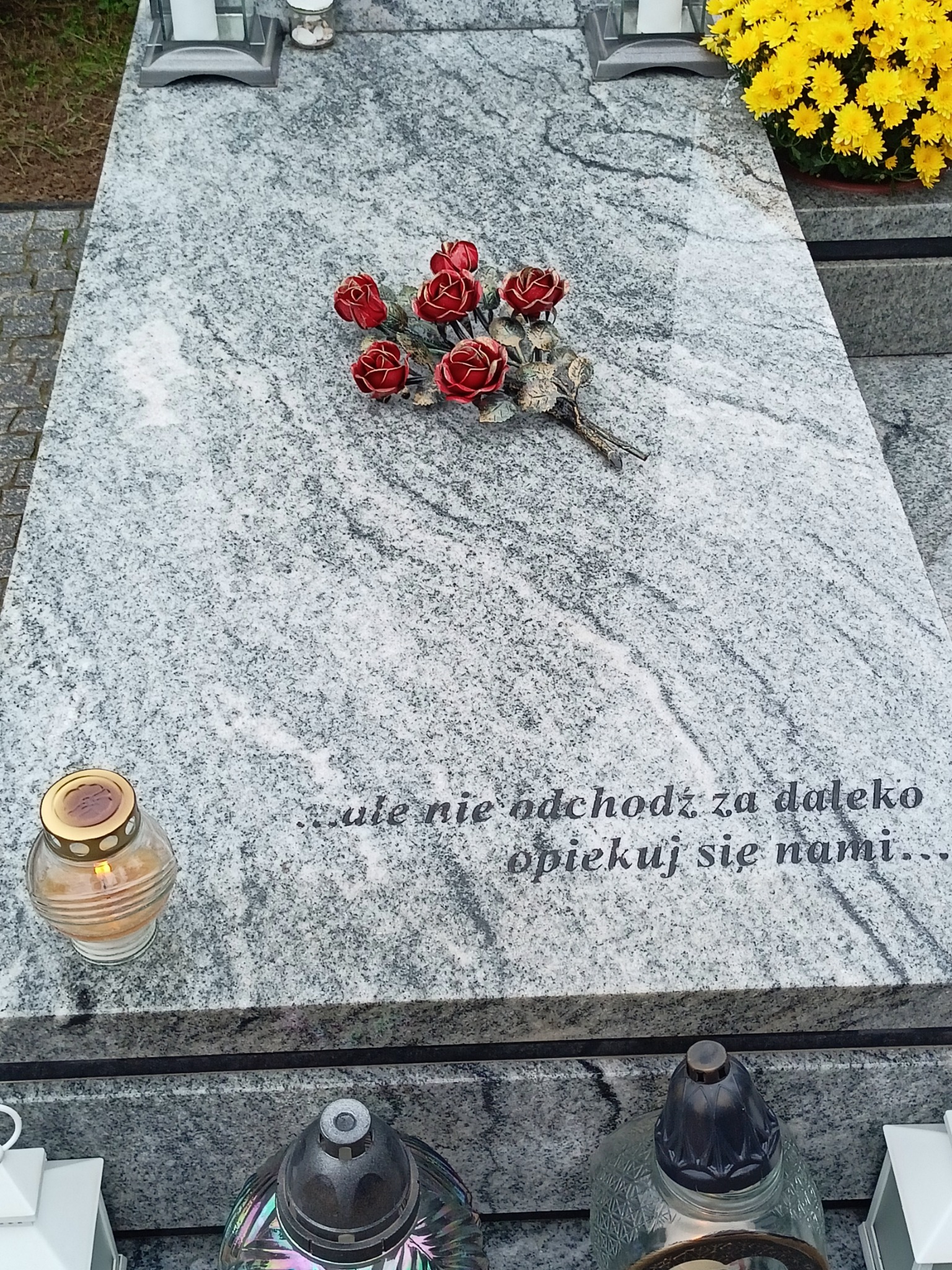 Granitowy nagrobek z wygrawerowanym napisem '...ule nie odchodź za daleko opiekuj się nami...', ozdobiony bukietem czerwonych róż z metalu i lampionem ze świecą, obok donica z żółtymi chryzantemami.