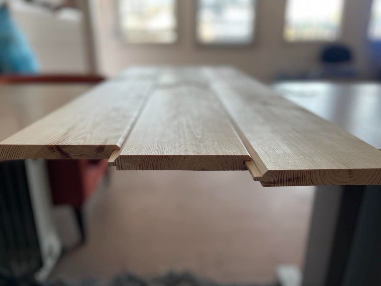 Profilowane deski boazeryjne z jasnego drewna, widoczne połączenia pióro-wpust, ułożone na stole w jasnym, rozproszonym świetle.