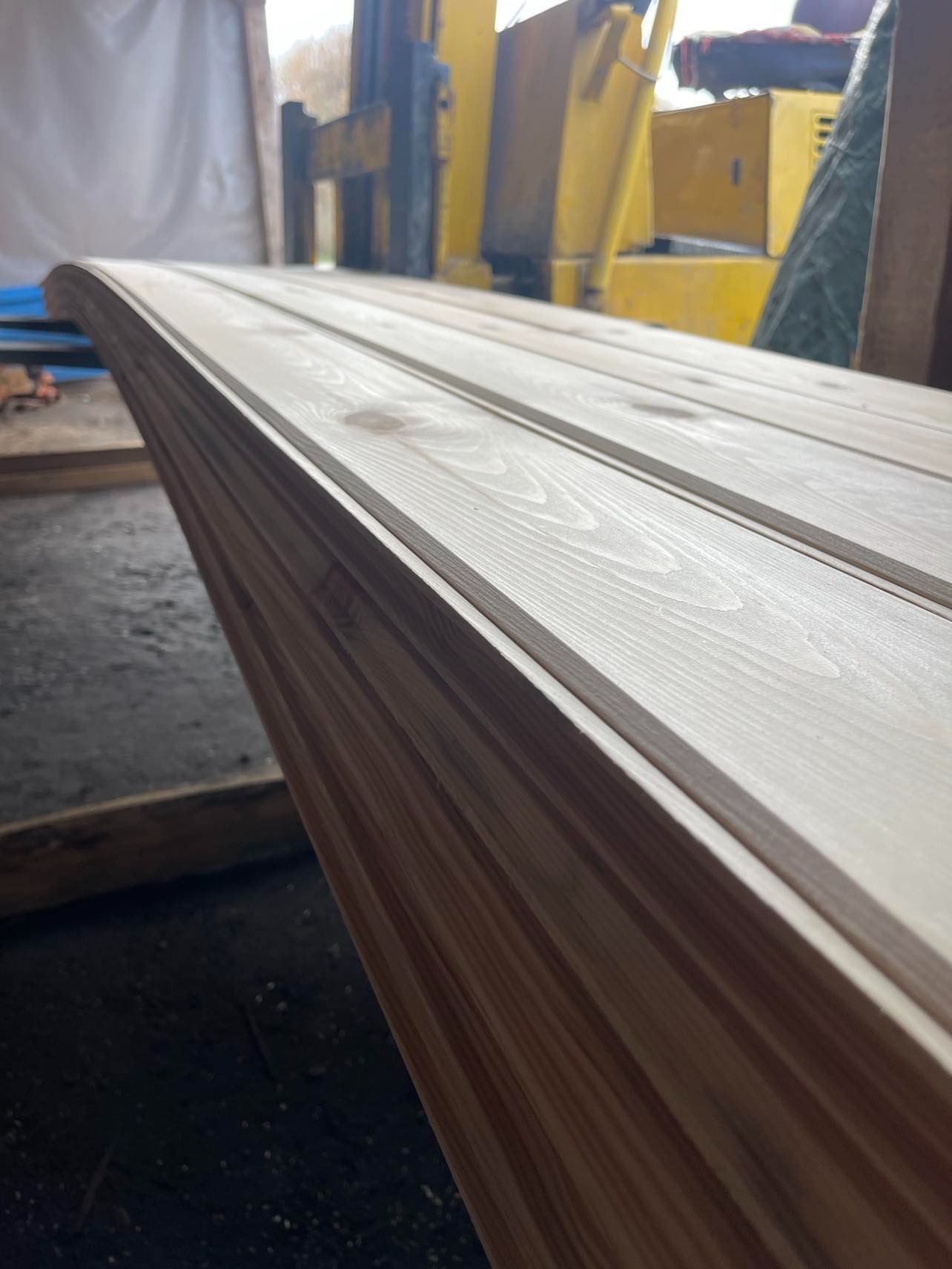 Stos drewnianych paneli z zaokrąglonymi krawędziami, przygotowanych do montażu, z żółtym wózkiem widłowym w tle.