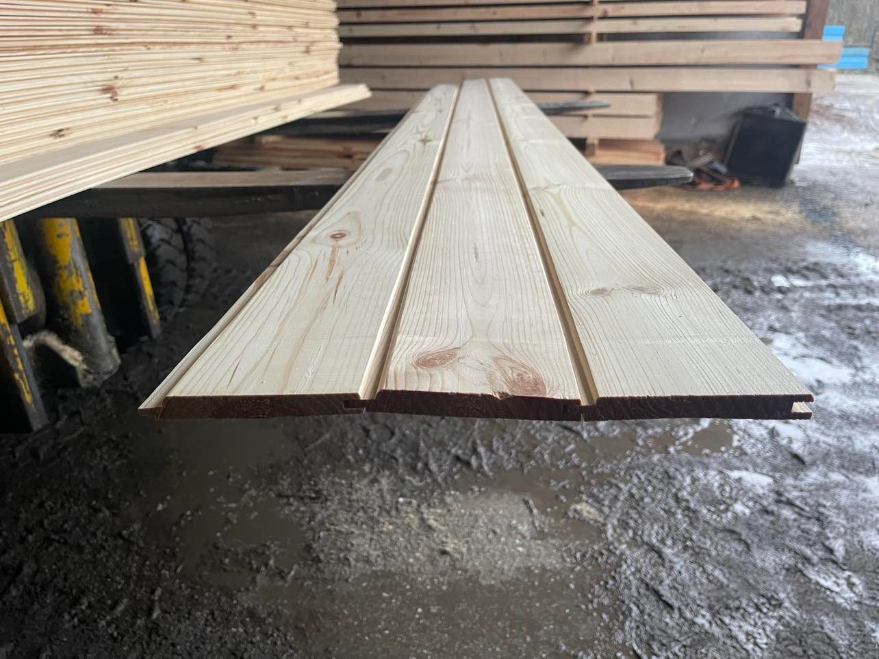 Profilowane deski boazeryjne z jasnego drewna, ułożone na tle stosu desek i błotnistego podłoża.