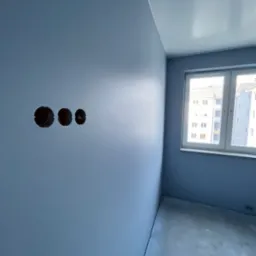 Ściana w odcieniach niebieskiego po świeżym malowaniu z trzema otworami pod gniazdka elektryczne, widoczne okno z widokiem na budynek mieszkalny.