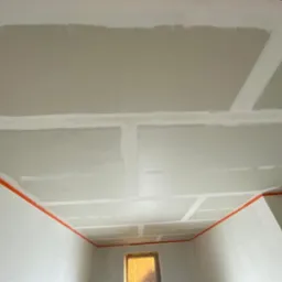 Sufit i ściany w trakcie prac wykończeniowych, widoczne łączenia płyt kartonowo-gipsowych wzmocnione taśmą, zabezpieczone krawędzie pomarańczową taśmą malarską.