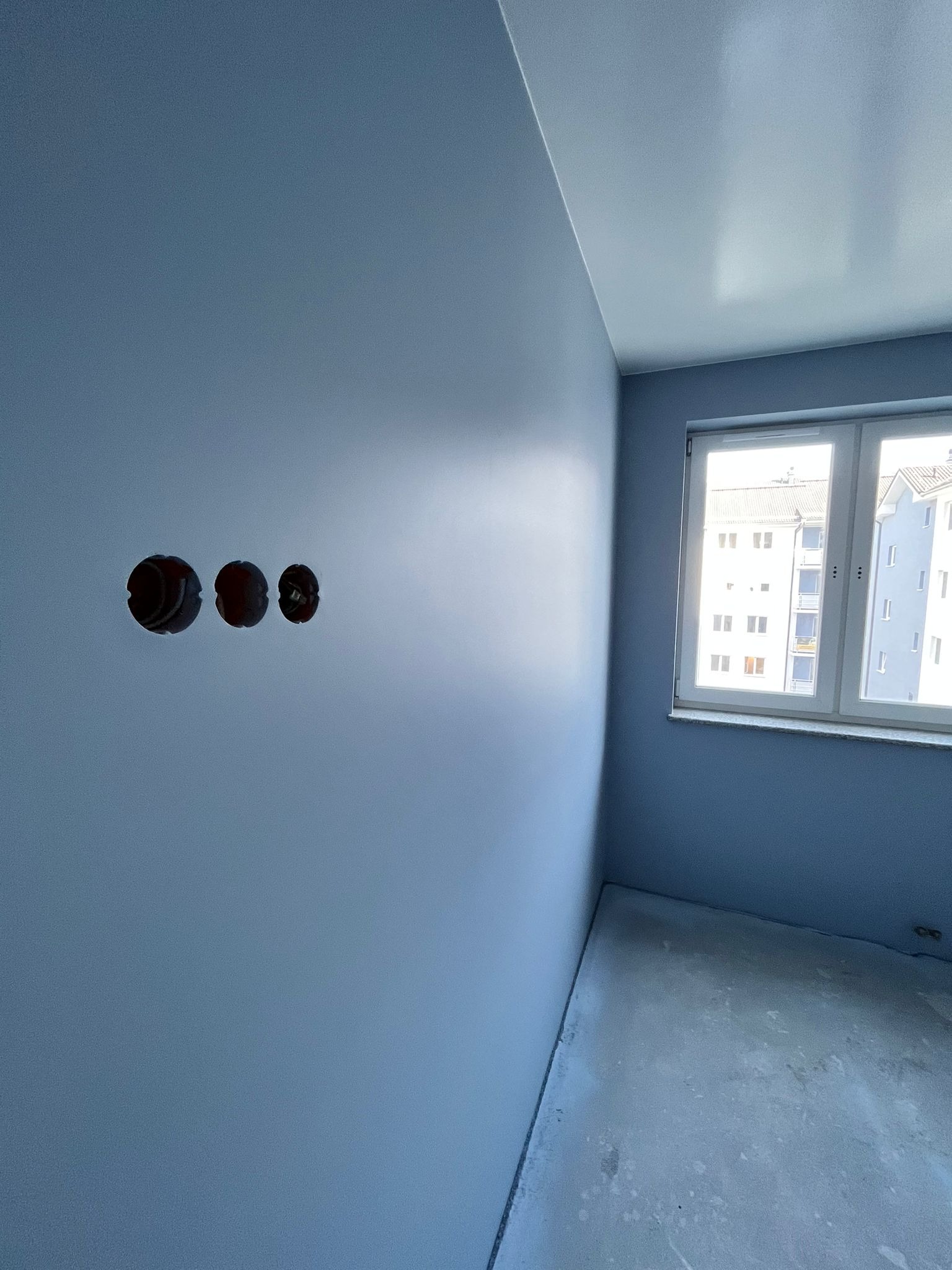 Ściana w odcieniach niebieskiego po świeżym malowaniu z trzema otworami pod gniazdka elektryczne, widoczne okno z widokiem na budynek mieszkalny.