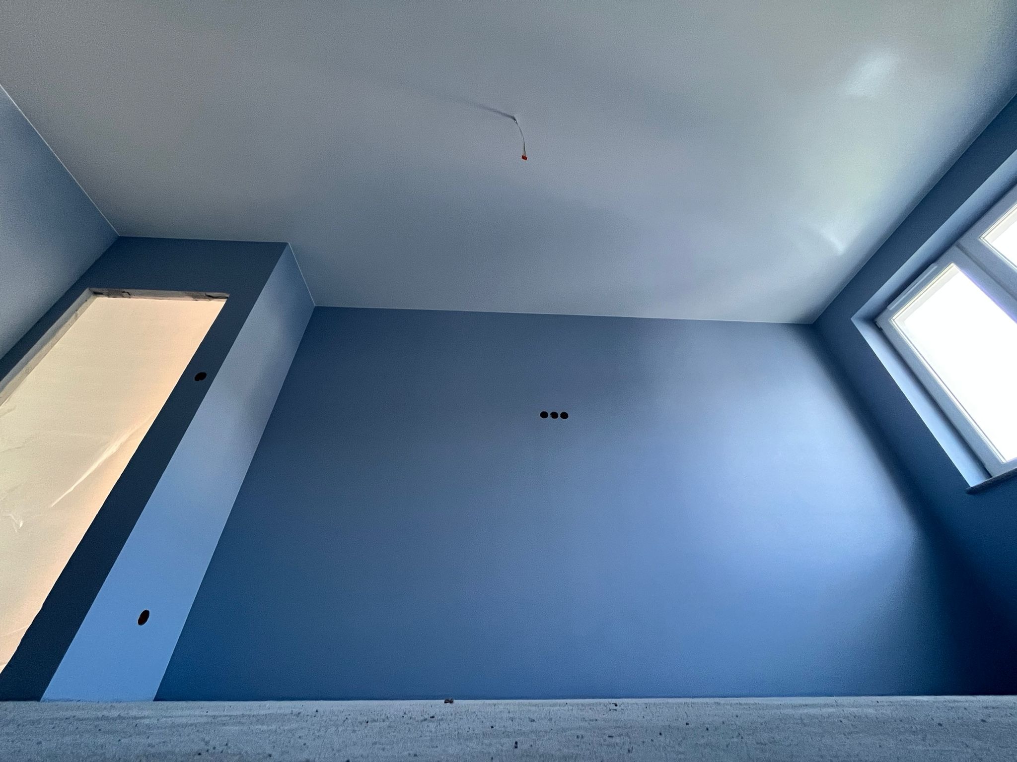 Świeżo pomalowane na niebiesko wnętrze z oknem i otworami na kable w suficie, widok z dołu na surową podłogę.