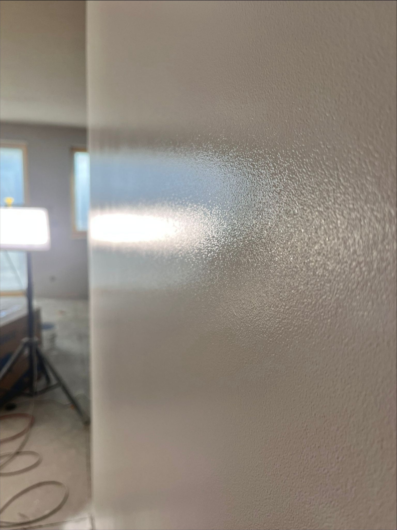 Świeżo pomalowana, gładka ściana w odcieniach beżu, z widoczną fakturą farby. W tle widoczne oświetlenie budowlane na statywie oraz fragment okna, sugerujące trwający remont.