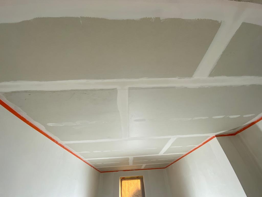 Sufit i ściany w trakcie prac wykończeniowych, widoczne łączenia płyt kartonowo-gipsowych wzmocnione taśmą, zabezpieczone krawędzie pomarańczową taśmą malarską.