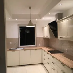 Jasna kuchnia w stylu skandynawskim z kremowymi szafkami, drewnianym blatem i beżowymi płytkami, oświetlona wiszącą lampą i halogenami.