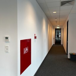 Wojciech Szymończyk KNS Complex - Korytarz biurowy z białymi ścianami, szarą wykładziną i czerwonym hydrantem na ścianie.