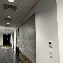 Wojciech Szymończyk KNS Complex - Korytarz biurowy przygotowany do malowania: podłoga i szklane elementy zabezpieczone folią, ściany świeżo pomalowane na biało, widoczne taśmy malarskie pod sufitem.