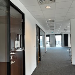 Wojciech Szymończyk KNS Complex - Wnętrze biura z szarą wykładziną, białymi ścianami i czarnymi ramami szklanych drzwi, widok na korytarz z oknami.