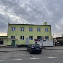 Dwukondygnacyjny budynek biurowy w odcieniach zieleni i beżu, z zaparkowanym czarnym samochodem na pierwszym planie i paczkomatem InPost obok.