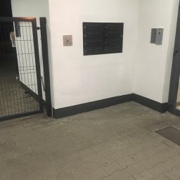 Wojciech Szymończyk KNS Complex - Biała elewacja budynku z czarnymi skrzynkami na listy, domofonem i metalową bramą wejściową, widok z zewnątrz.