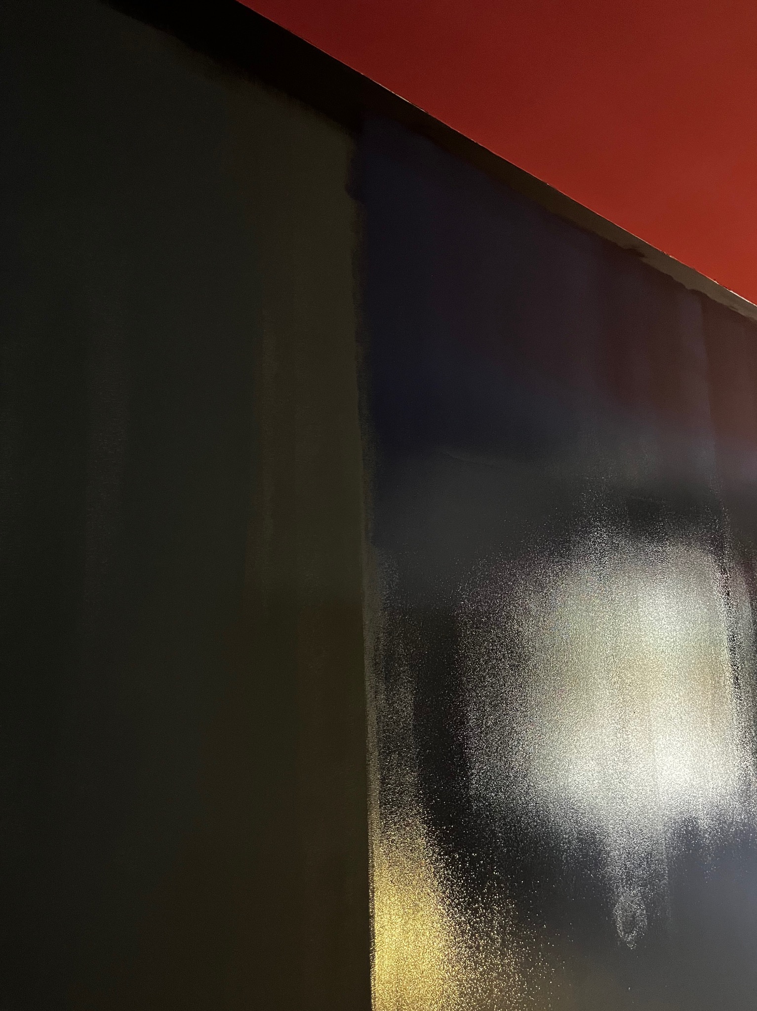 Świeżo pomalowana ściana na czarno, z widocznym połyskiem i fragmentem czerwonego sufitu w rogu.