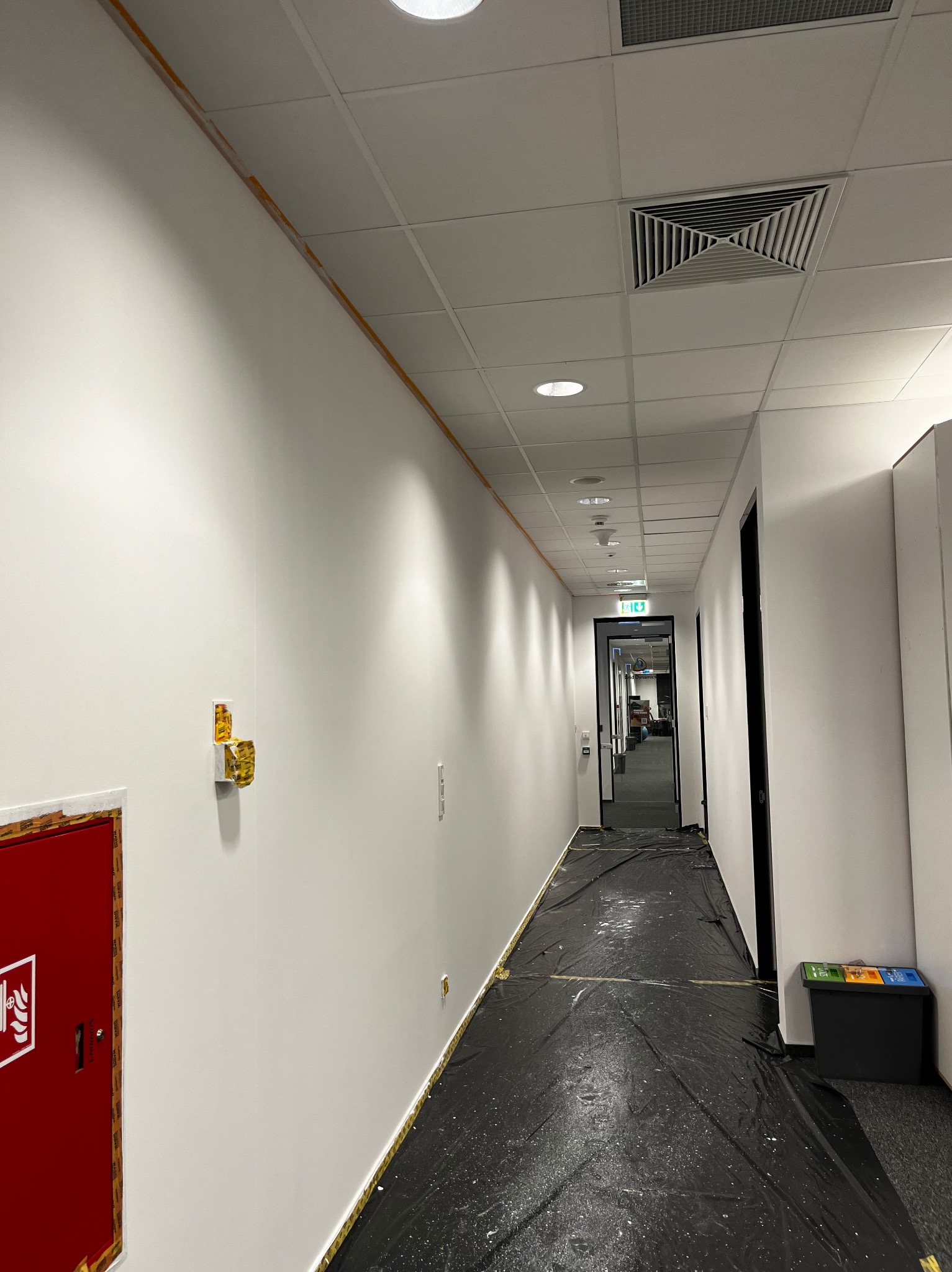 Korytarz biurowy w trakcie odświeżania: ściany zabezpieczone taśmą malarską, podłoga przykryta folią ochronną, widoczna skrzynka hydrantowa i wentylacja sufitowa.