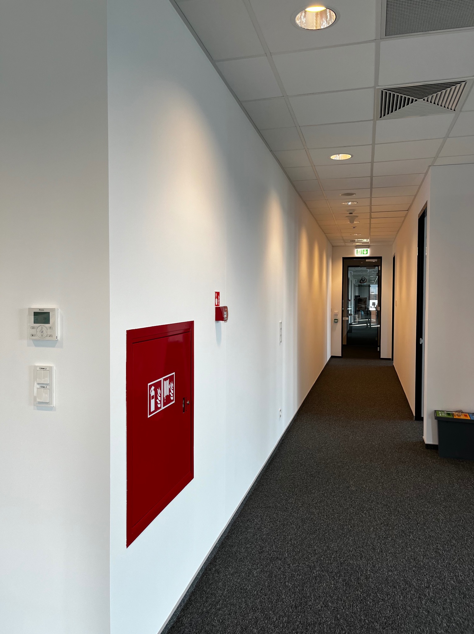 Korytarz biurowy z białymi ścianami, szarą wykładziną i czerwonym hydrantem na ścianie.