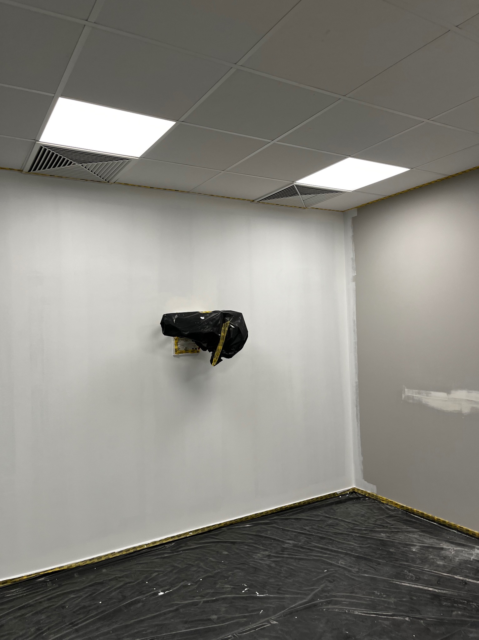 Przygotowanie do malowania biura w Smolcu: ściany zabezpieczone folią i taśmą malarską, gniazdko elektryczne zakryte folią ochronną, widoczny fragment świeżo pomalowanej ściany.