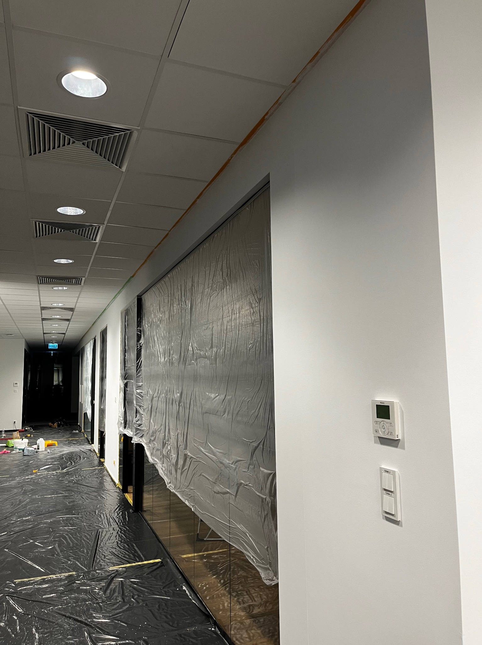 Korytarz biurowy przygotowany do malowania: podłoga i szklane elementy zabezpieczone folią, ściany świeżo pomalowane na biało, widoczne taśmy malarskie pod sufitem.